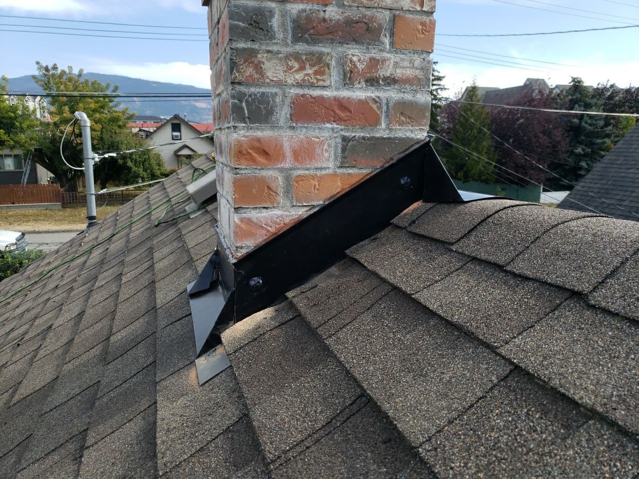 Black sheet-metal cricket on the uphill side of a brick chimney on a shingle roof