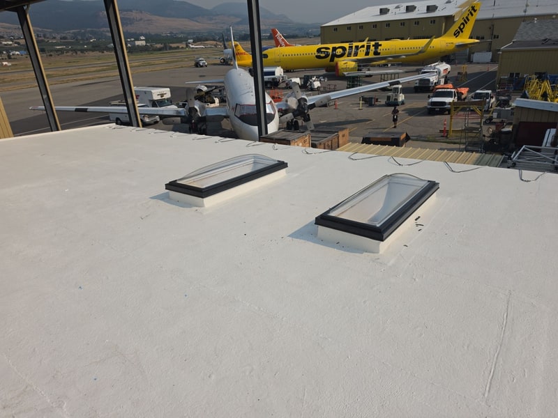 Two curb-mounted skylights on a white flat roof, aircraft parked on the apron beyond