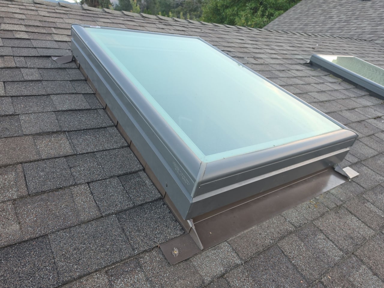 Grey-framed skylight on a shingle roof, metal flashing along its sides, a second skylight behind it