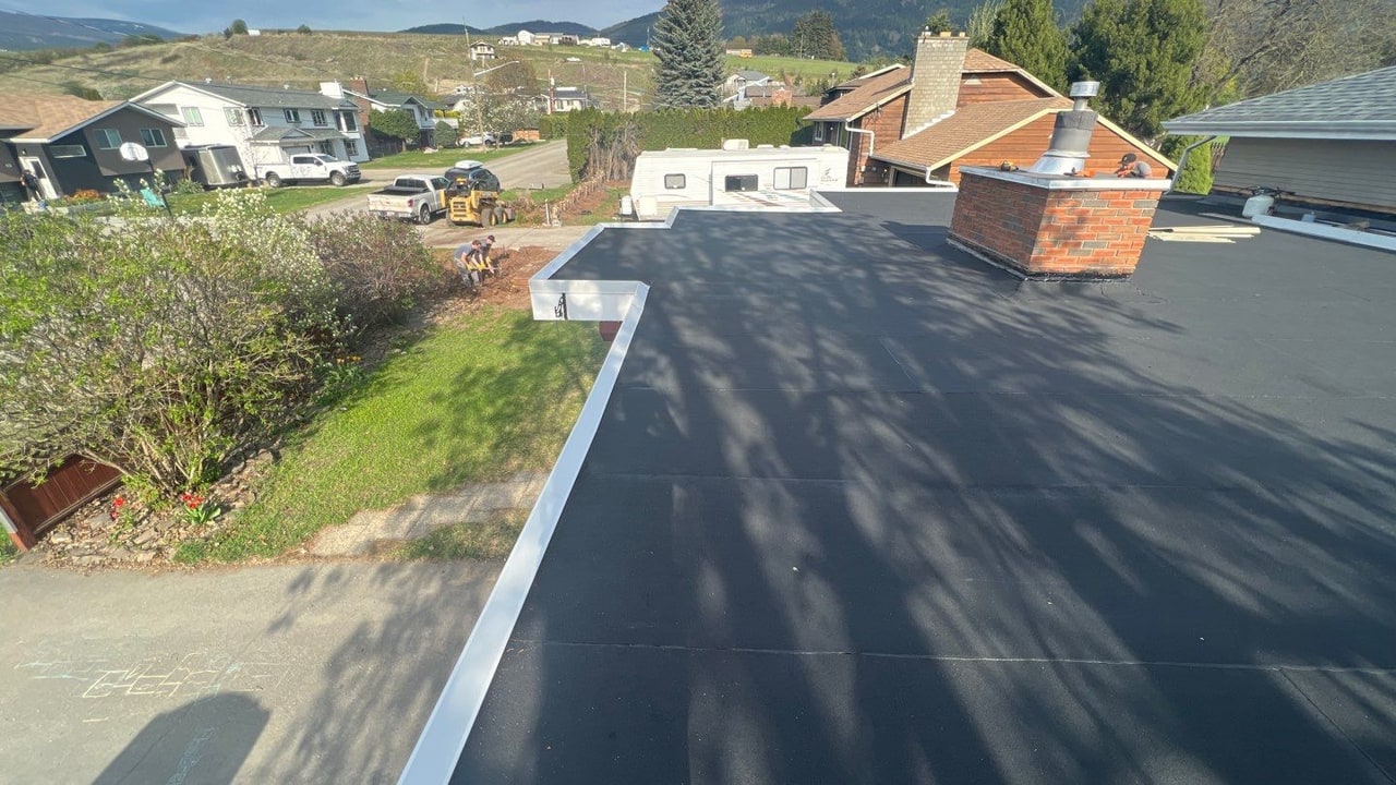 New black torch-on flat roof with white edge metal and a re-flashed brick chimney, the hillside behind